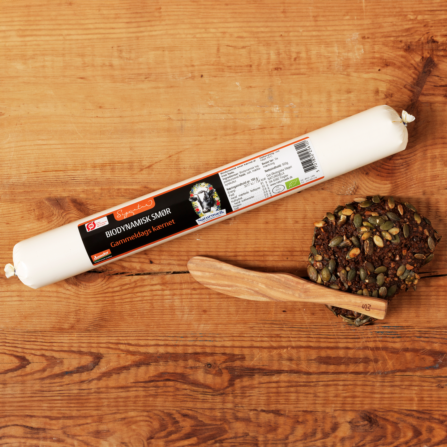 Biodynamisk smør på træbord ved siden af fuldkornsrundel med kerner og trækniv.