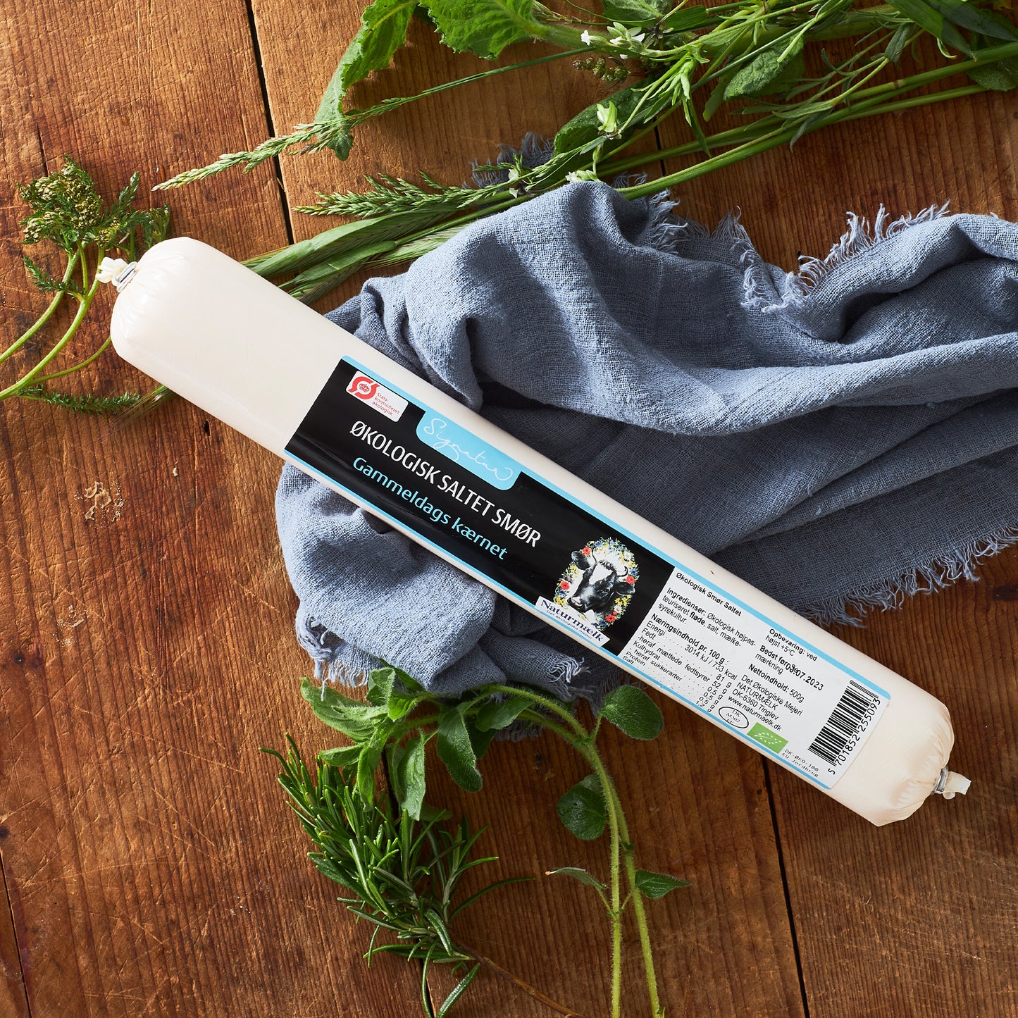 Rulle af økologisk saltet smør på træbord med blå stof og friske urter
